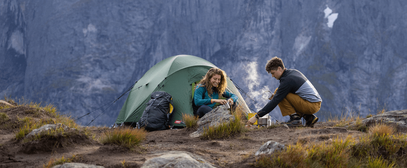 Two people cooking outside a green tent on a rocky ridge with massive mountains behind.